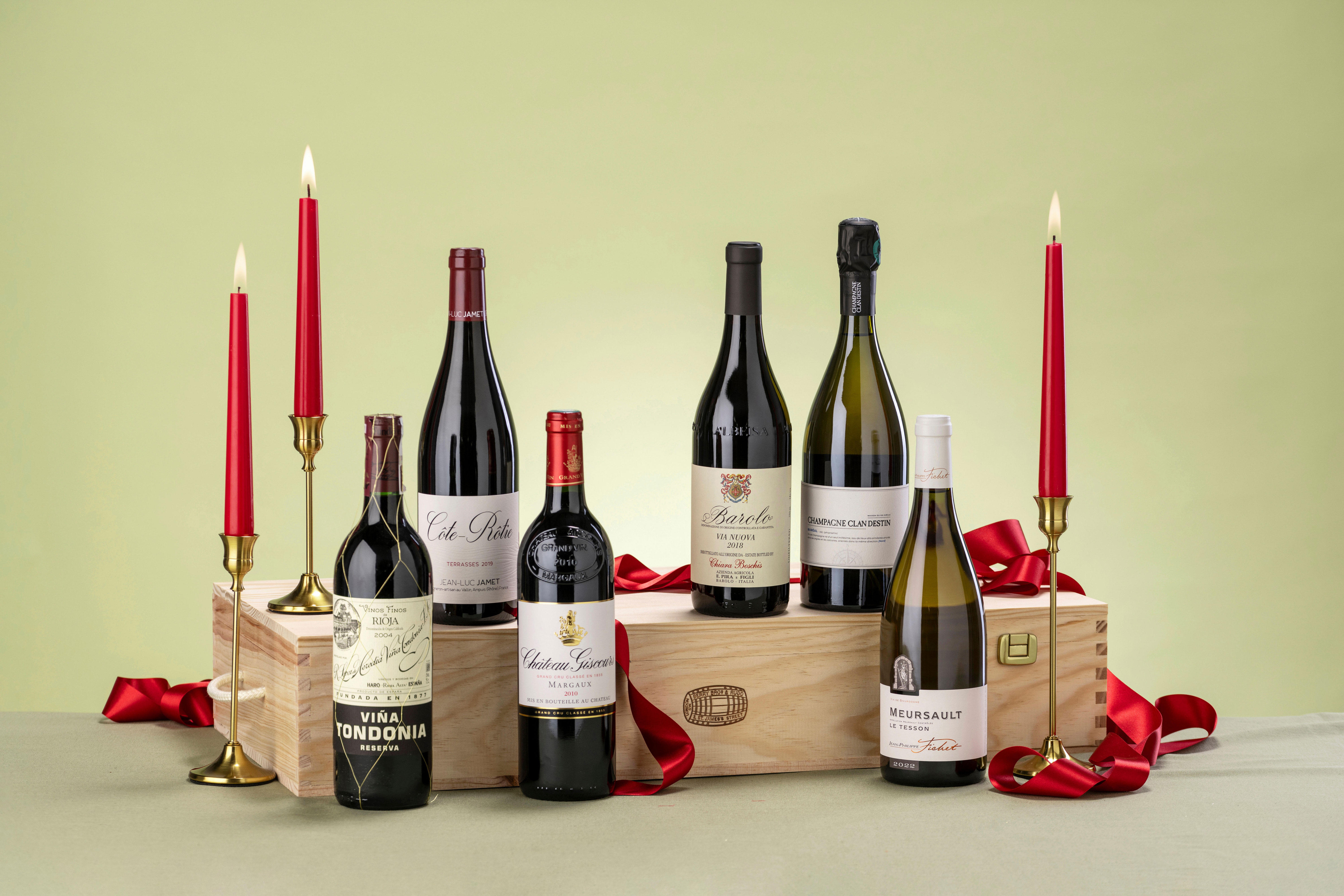 Berry Bros. & Rudd gifting set displayed against a soft green background. The set includes a bottle of Champagne Leclerc Briant with a gold foil top, a tall brown gift box, and a tin of Marc de Champagne truffles with some truffles visible. Festive accents include a red ribbon, a lit red candle in a brass holder, and a small wooden Christmas tree, creating an elegant seasonal presentation.