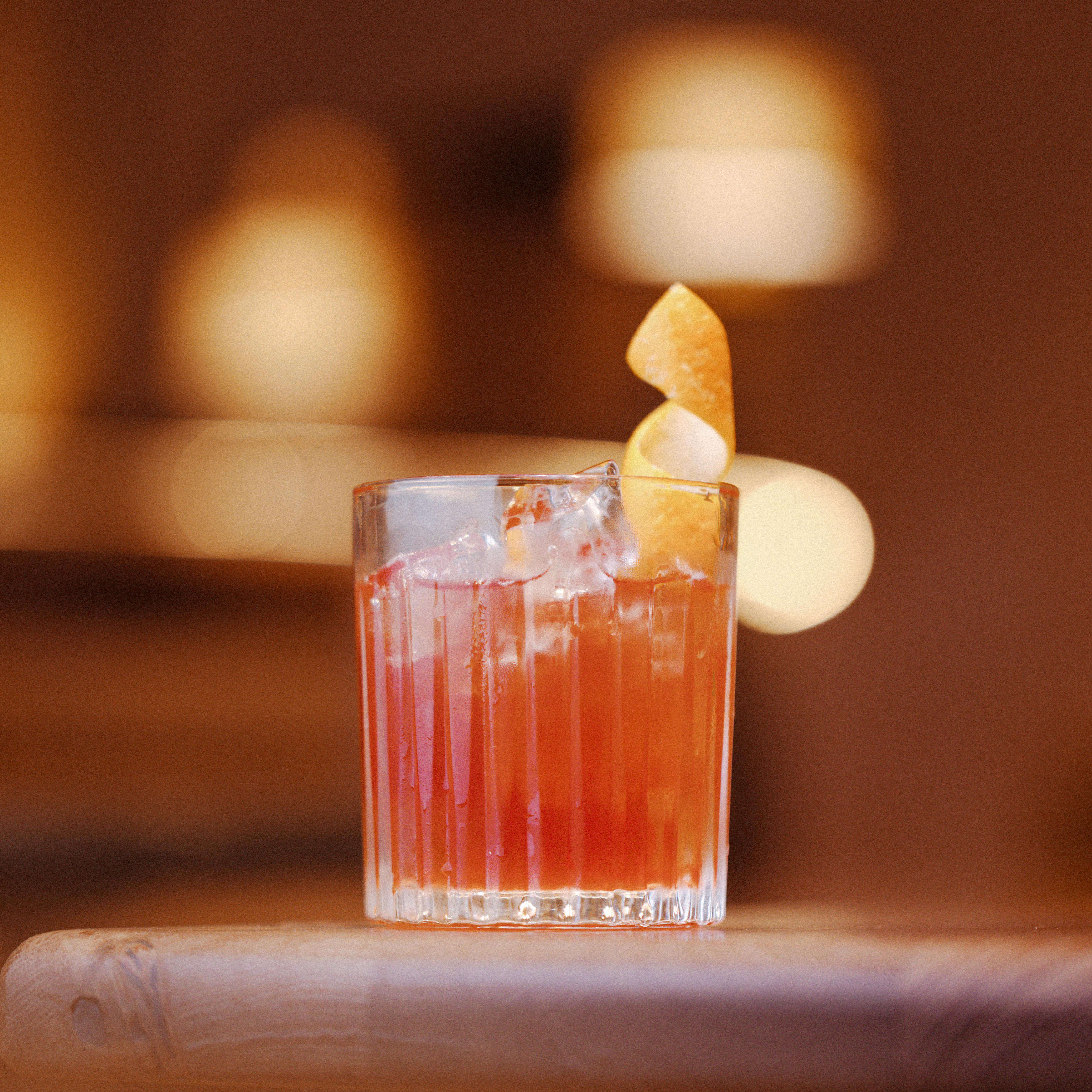 A Negroni-style cocktail served over ice in a cut-glass tumbler, garnished with an orange twist, and set against a warm, softly lit background — part of the Berry Bros. & Rudd cocktail range.