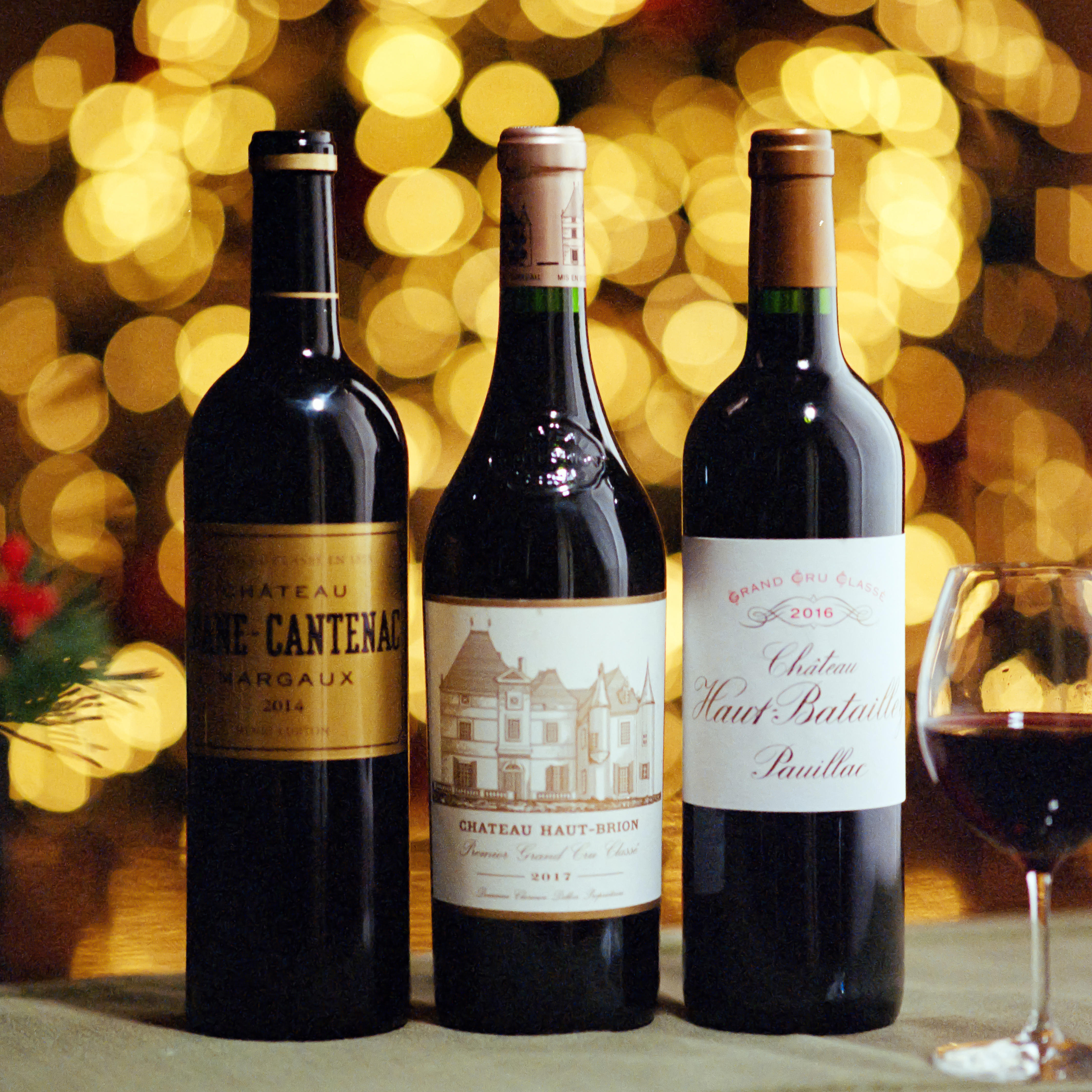 Three bottles of fine Bordeaux claret on a festive table at Berry Bros & Rudd: Château Brane-Cantenac Margaux, Château Haut-Brion, and Château Haut-Batailley, with warm golden bokeh lights in the background.