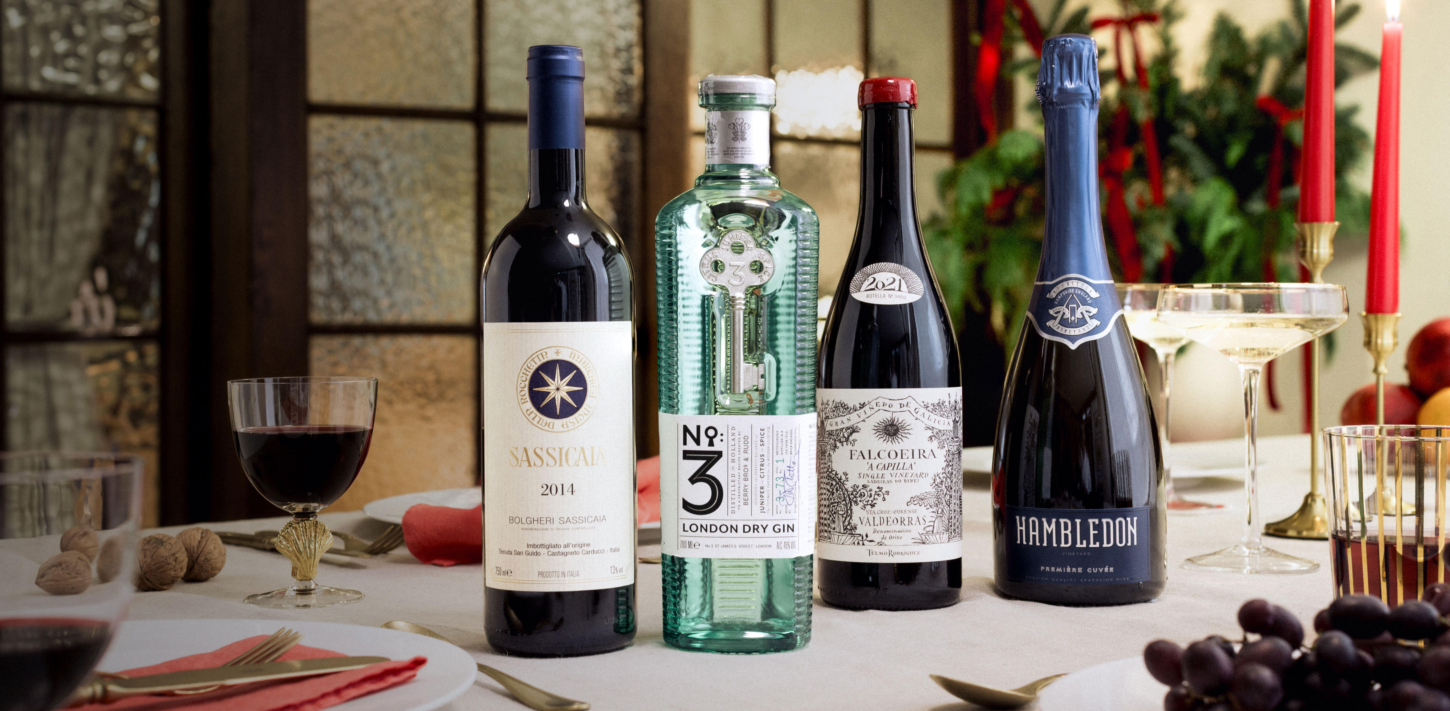 Festive dining table with four bottles: Sassicaia red wine, No.3 London Dry Gin, Château Talbot red wine, and Hambledon sparkling wine, surrounded by glasses, grapes, nuts, and red candles in a Christmas setting.