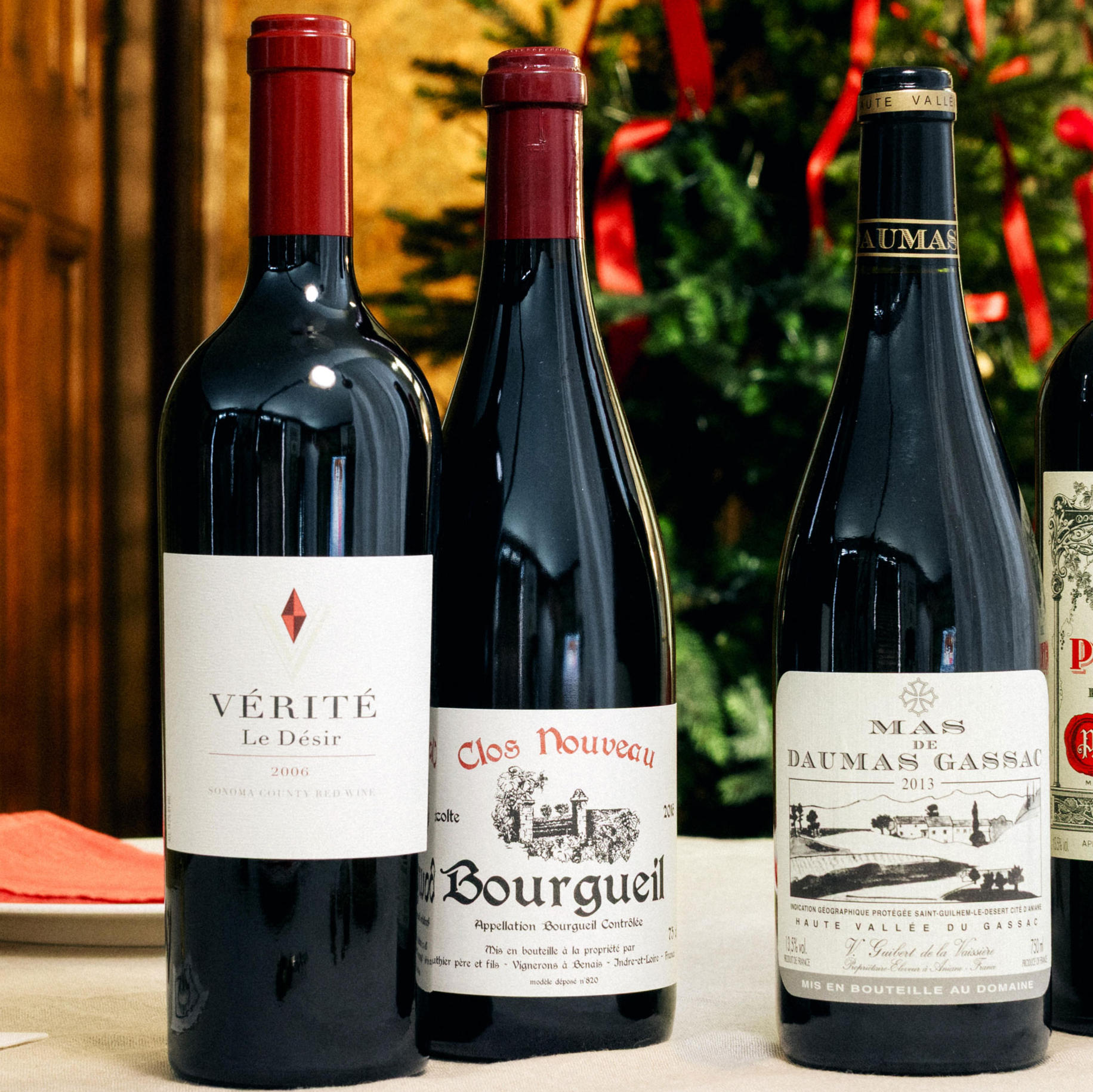 Three bottles of mature fine wine are displayed on a festive table with a Christmas tree in the background. The bottles include Vérité Le Désir 2004, Clos Nouveau Bourgueil, and Mas de Daumas Gassac 2013, each featuring distinctive labels and deep red or black foil tops. Seasonal decorations and warm holiday lighting complete the elegant setting.