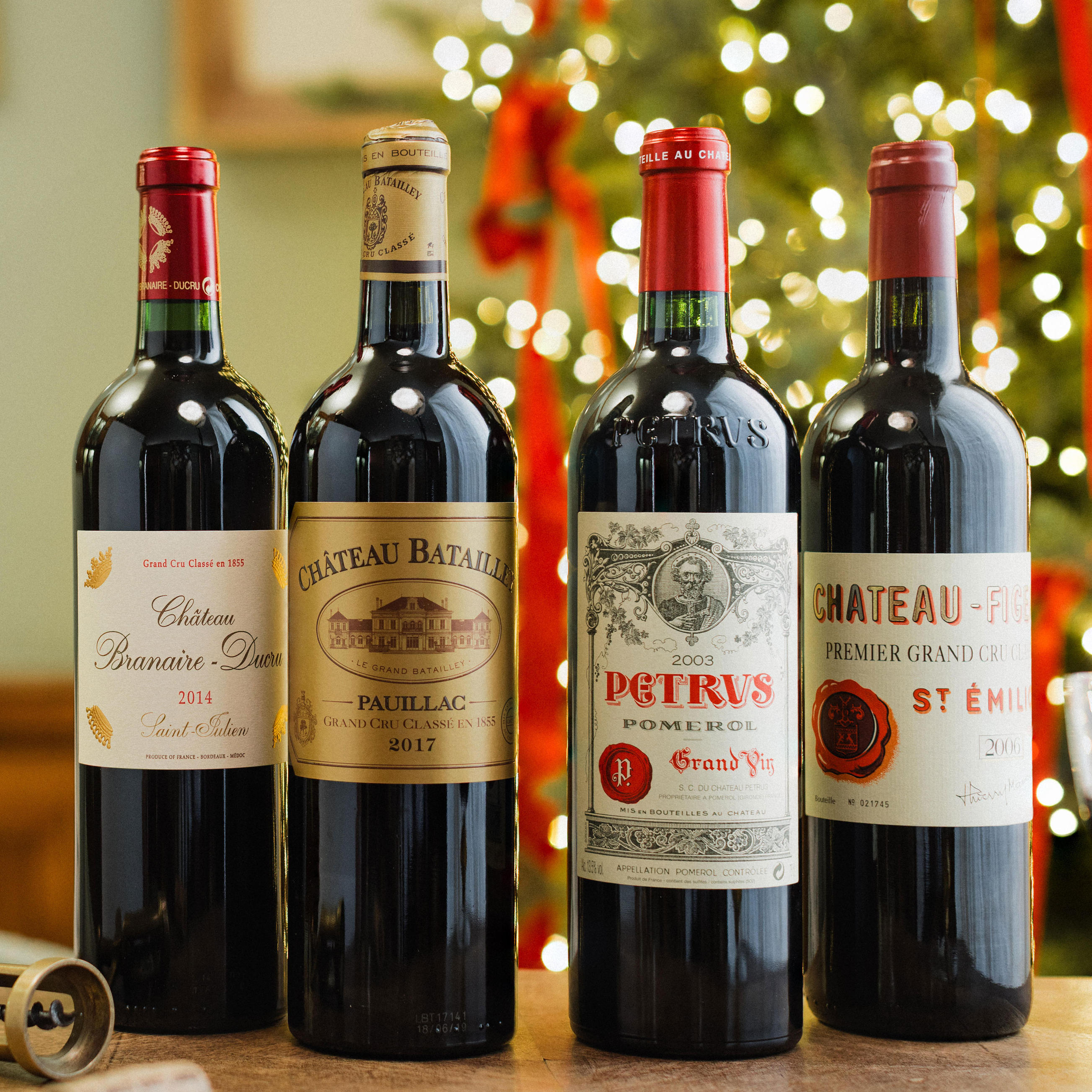 Four bottles of fine Claret are displayed in front of a Christmas tree with warm festive lights. The bottles include Château Branaire-Ducru 2014, Château Batailley Pauillac 2017, Petrus Pomerol 2003, and Château Figeac St. Émilion. Each bottle features distinctive labels and red foil tops, arranged on a wooden surface for an elegant holiday presentation.