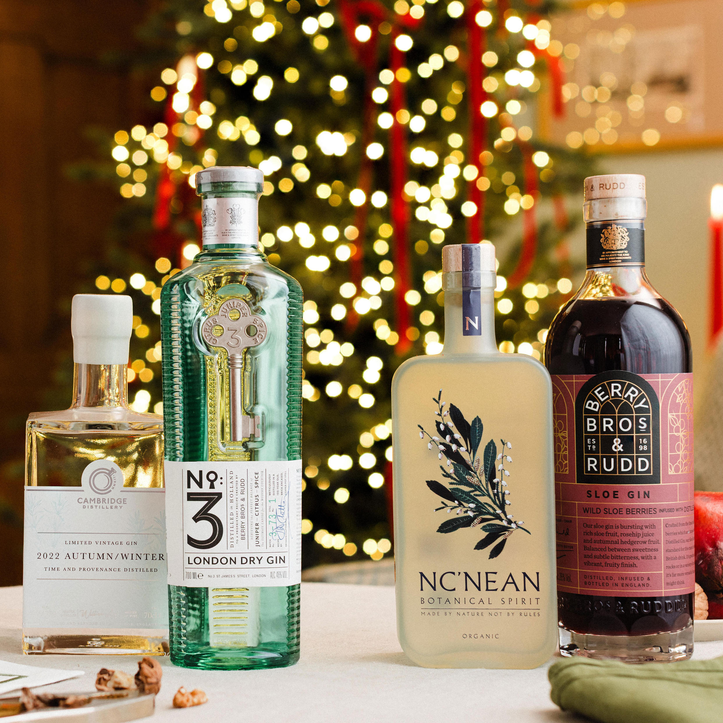 Four bottles of gin are displayed on a Christmas-themed table with a decorated tree and red candles in the background. The selection includes Cambridge Distillery gin, No.3 London Dry Gin in a green bottle, Nc’Nean Botanical Spirit in a frosted bottle, and Berry Bros. & Rudd Sloe Gin in a dark bottle with a pink label. Seasonal accents such as pomegranate and festive greenery complete the elegant holiday setting.
