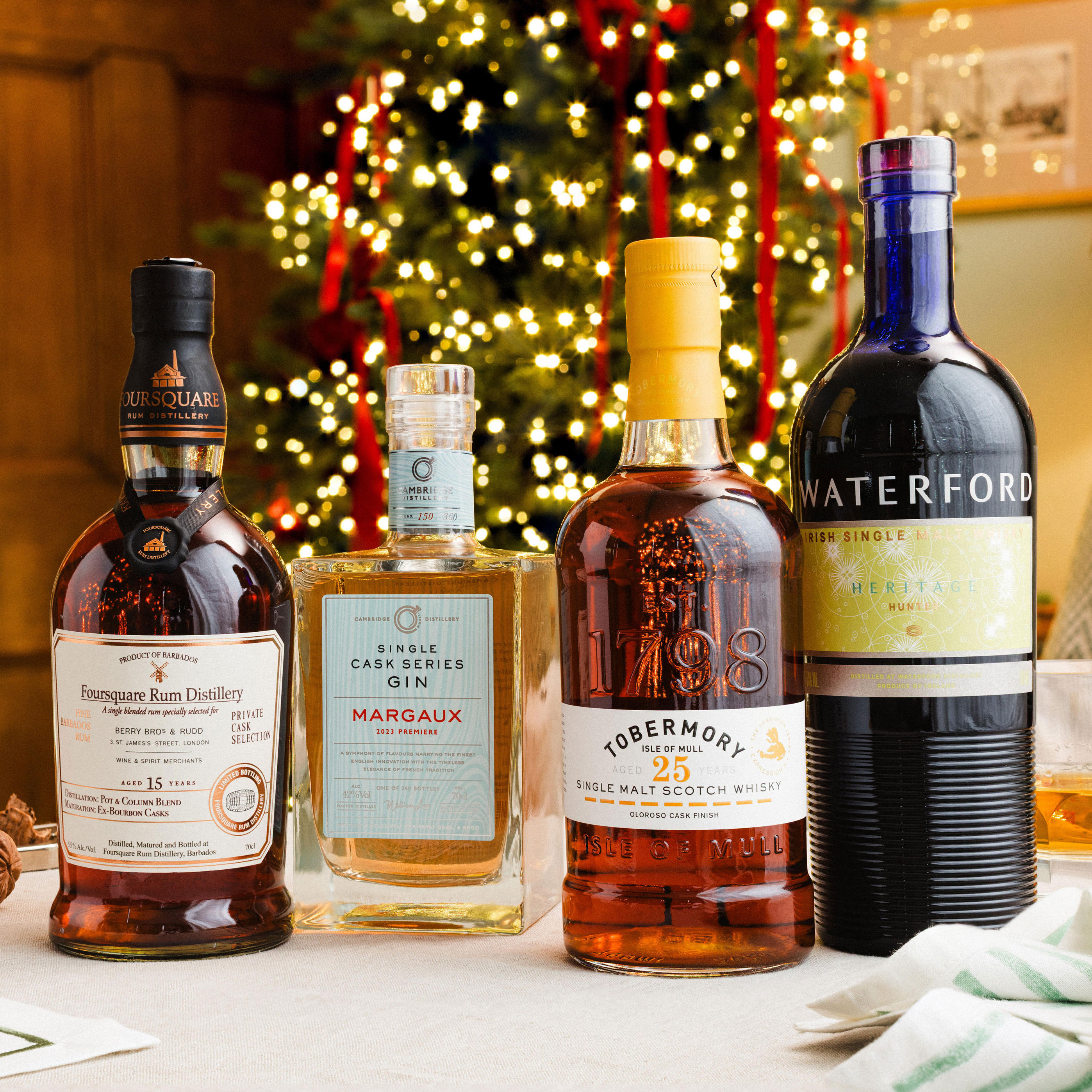 Four bottles of premium spirits displayed on a festive table with a Christmas tree and red ribbons in the background. The selection includes Foursquare Rum Distillery 15 Year Old, Cambridge Distillery Single Cask Series Gin Margaux, Tobermory 25 Year Old Single Malt Scotch Whisky, and Waterford Irish Single Malt Whisky. Seasonal accents such as nuts and warm holiday lighting complete the elegant setting.