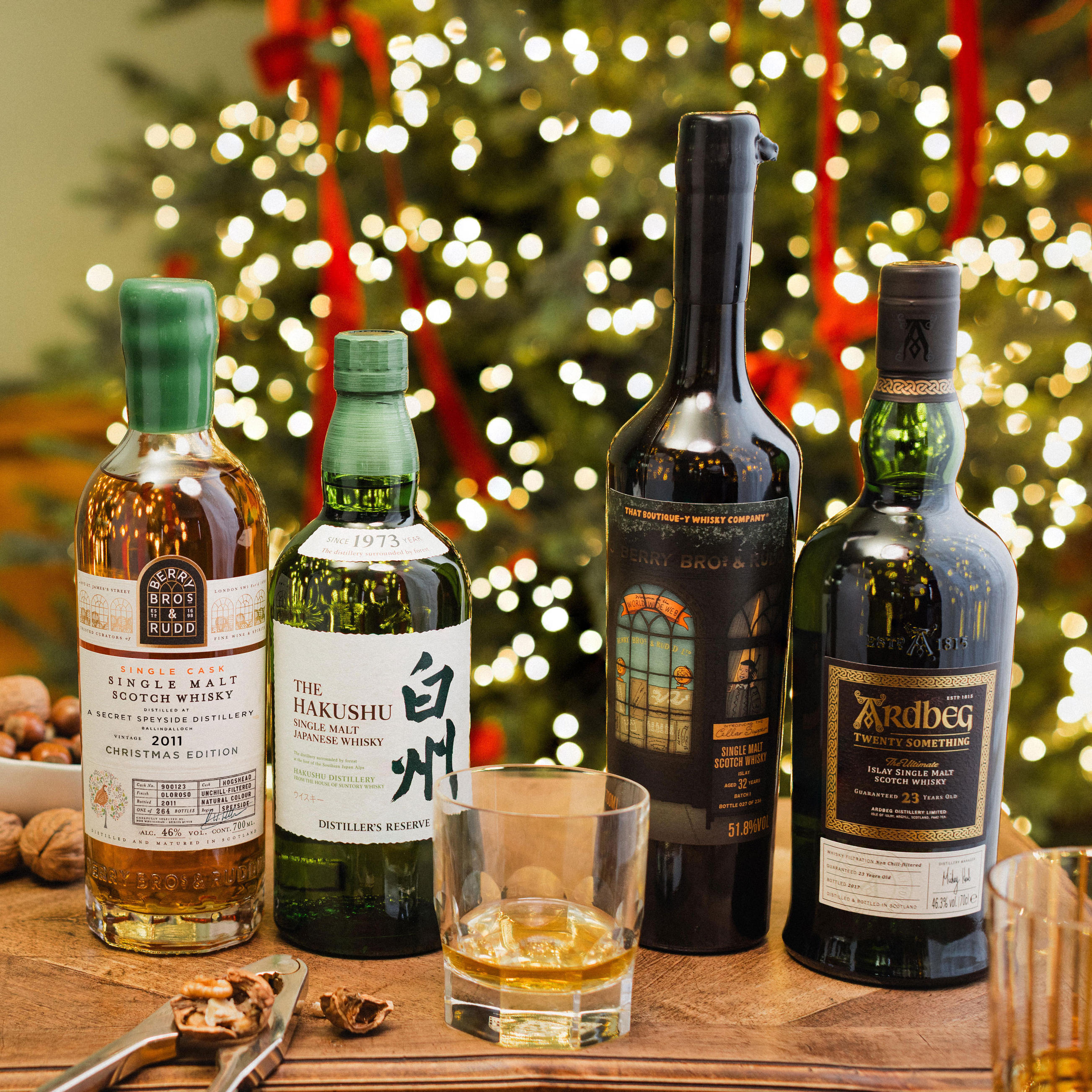 Four bottles of whisky are displayed on a wooden table with a Christmas tree and red ribbons in the background. The selection includes Berry Bros. & Rudd Single Malt Scotch Whisky 2011, The Hakushu Single Malt Japanese Whisky, Bruichladdich Single Malt Scotch Whisky, and Ardbeg Twenty Something Islay Single Malt Scotch Whisky. A glass of whisky and festive accents, such as nuts, complete the warm holiday setting.