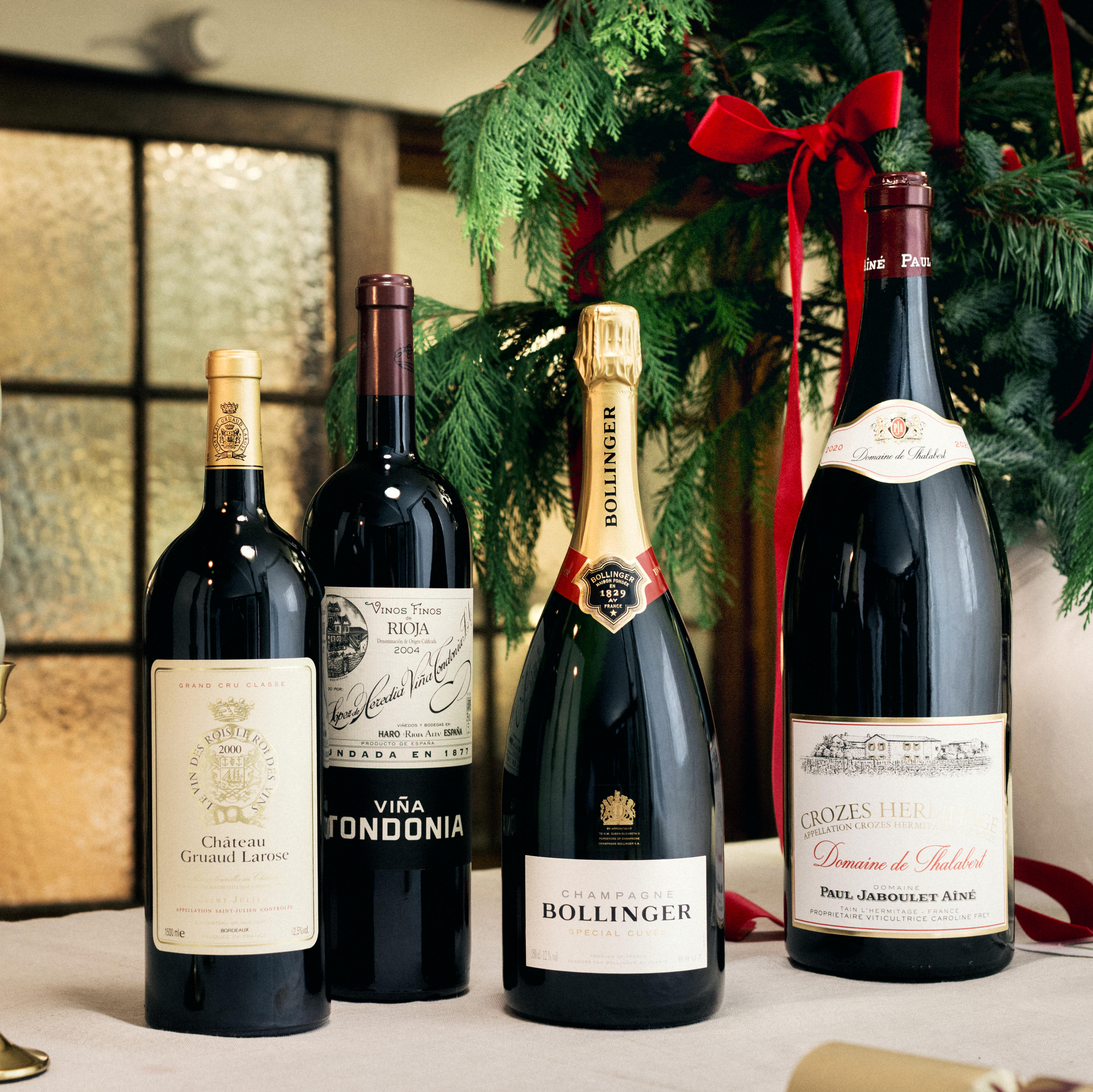 Four large-format bottles are displayed on a festive table, surrounded by greenery and red ribbons in the background. The selection includes Château Gruaud Larose, Viña Tondonia, Bollinger Champagne in a magnum bottle, and Crozes Hermitage Domaine de Thalabert. Each bottle features distinctive labels and foil tops, creating an impressive and celebratory holiday presentation.