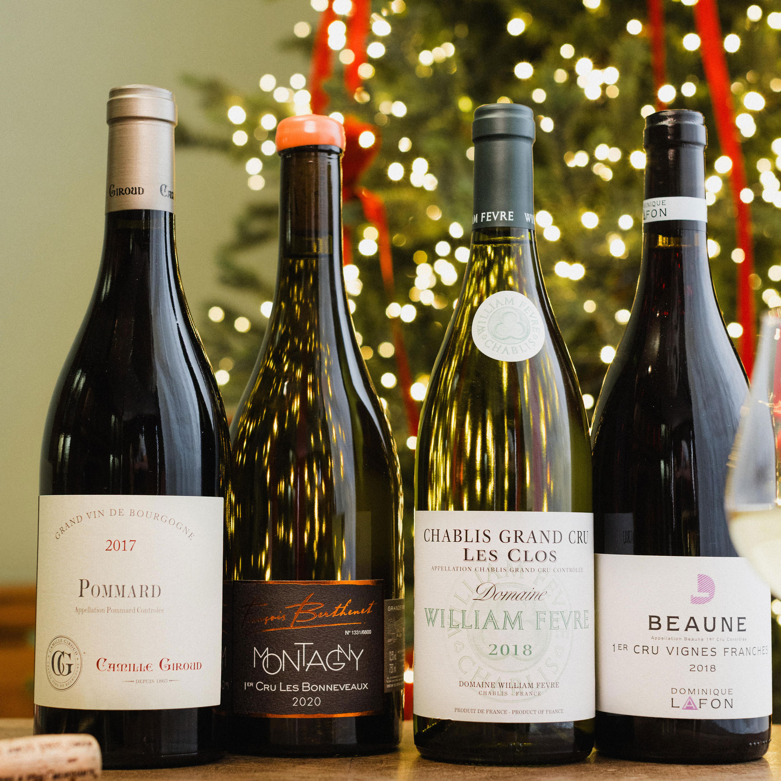 Four bottles of Burgundy wine are displayed in front of a Christmas tree with festive lights and red ribbons. The selection includes Pommard 2017, Montagny Premier Cru 2016, Chablis Grand Cru Les Clos 2018, and Beaune Premier Cru Vignes Franches 2018. A glass of white wine sits to the side, completing the elegant holiday setting.