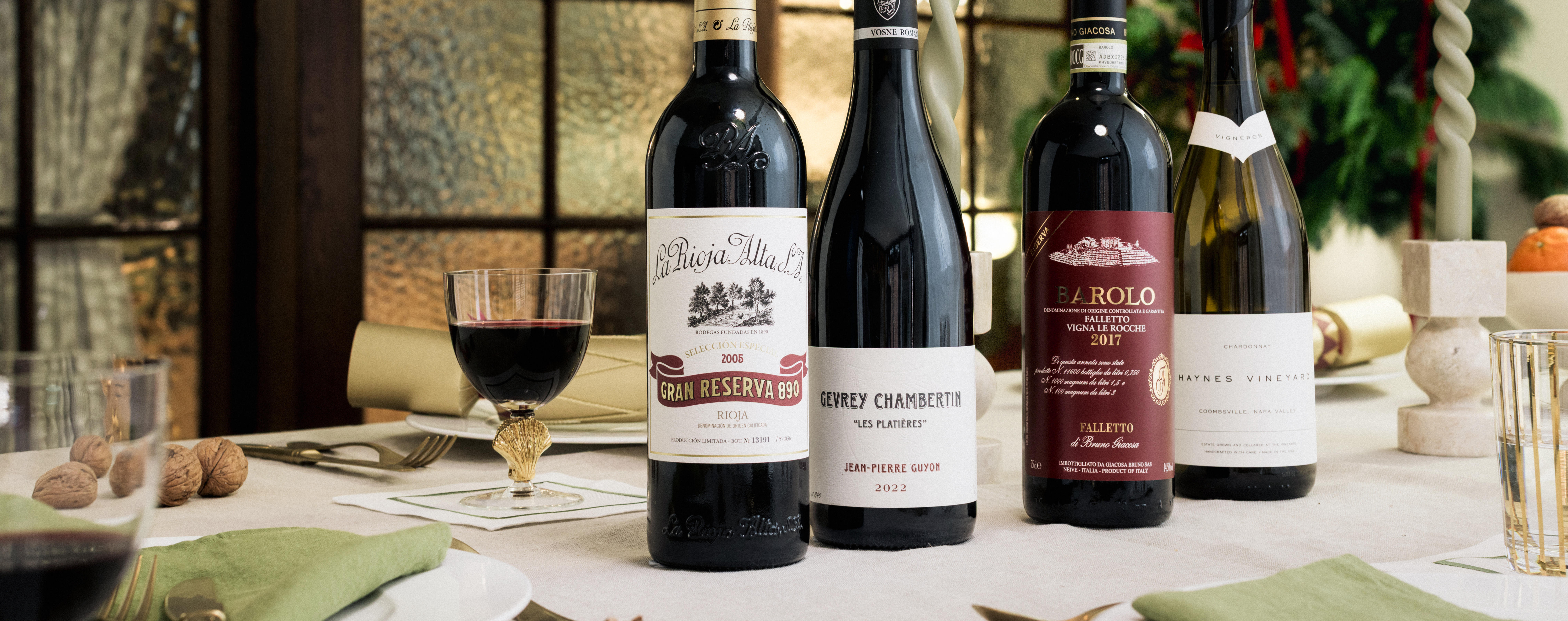 Four bottles of fine wine are displayed on an elegant Christmas table, surrounded by festive greenery and candles in the background. The selection includes La Rioja Alta Gran Reserva 890 2006, Gevrey-Chambertin “Les Evocelles” 2021, Barolo Riserva 2012, and a white wine from Domaine Roulot. Two glasses of red wine and seasonal decorations complete the luxurious holiday setting.