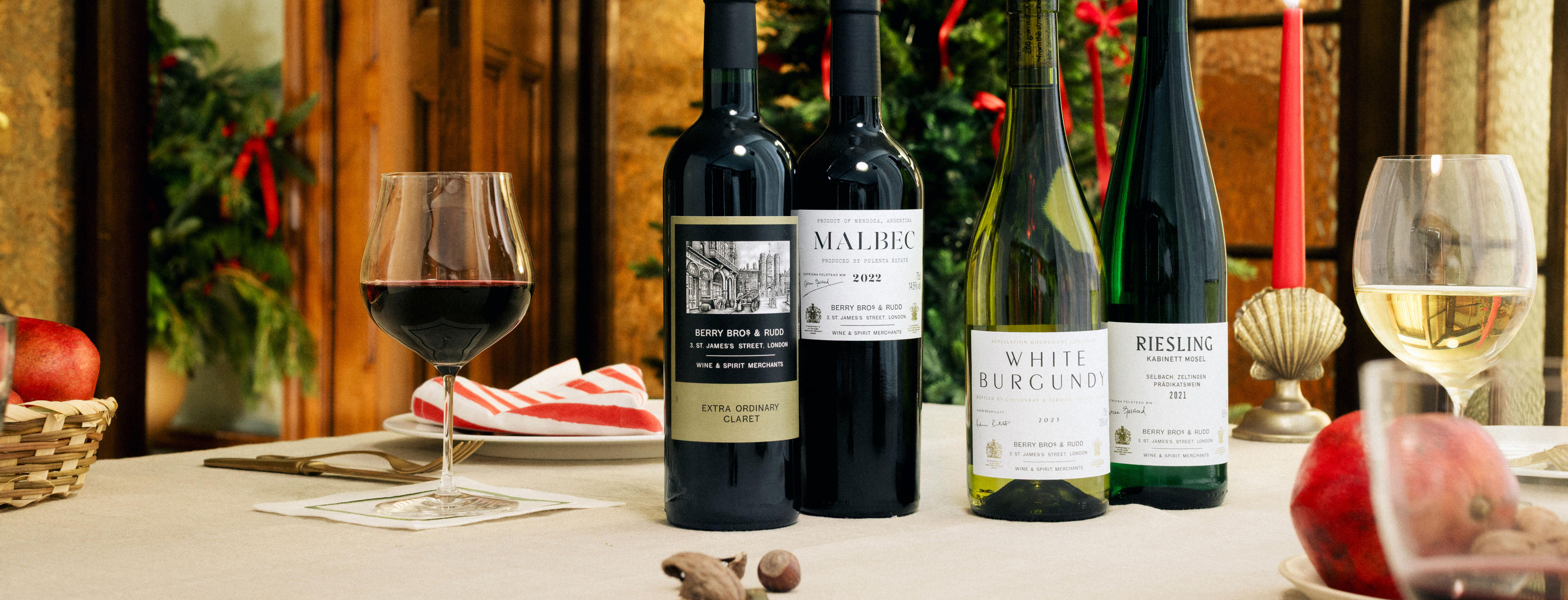 Four bottles from Berry Bros. & Rudd Own Selection range are displayed on a festive dining table. The wines include two reds (Berry Bros. & Rudd Claret and Malbec) and two whites (White Burgundy and Riesling). A glass of red wine and a glass of white wine sit alongside seasonal decorations, including pomegranates, nuts, and a red candle, with a Christmas tree in the background.