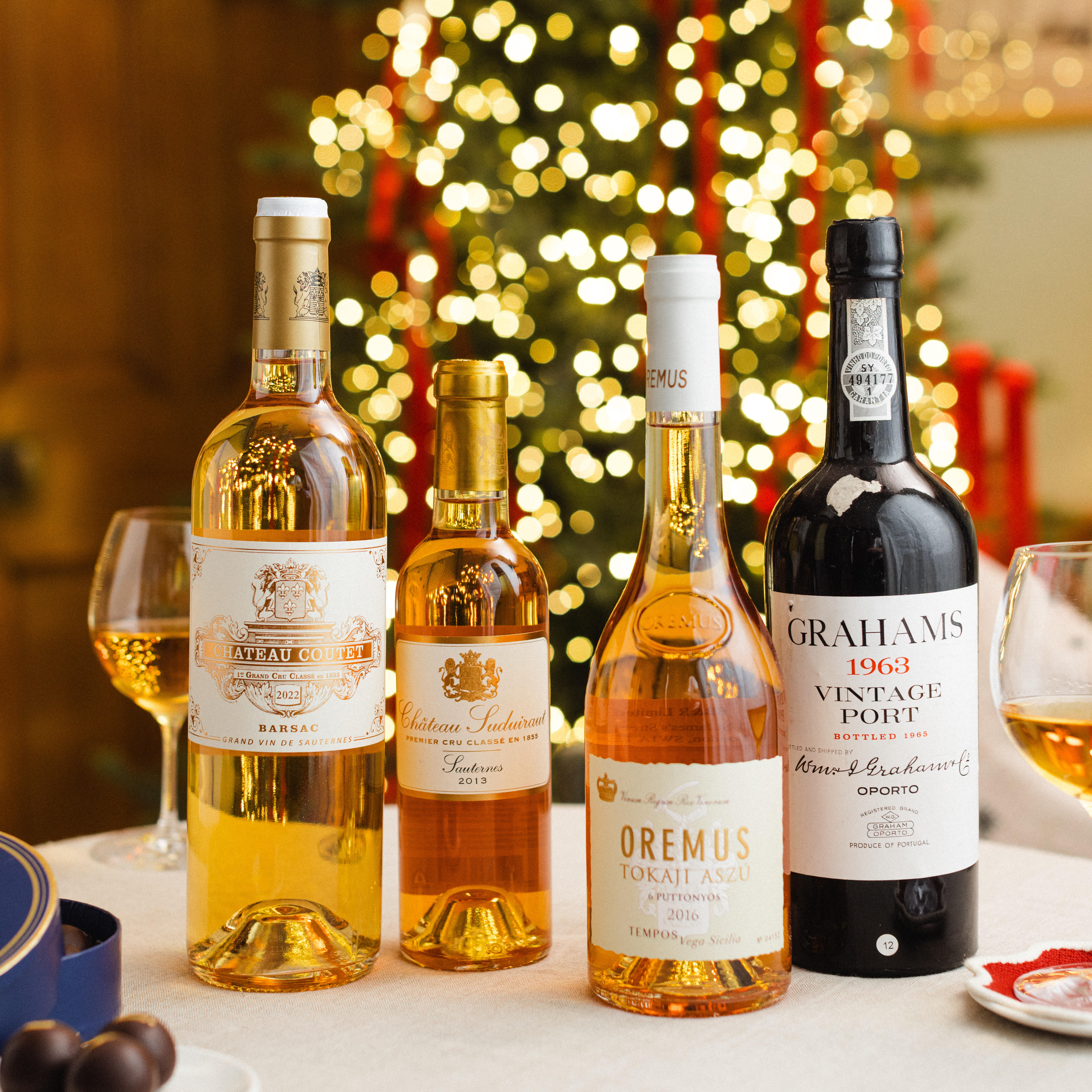 Four bottles of Port and dessert wine are displayed on a festive table, accompanied by a Christmas tree and warm lights in the background. The selection includes Château Coutet Barsac, Château Suduiraut Sauternes, Oremus Tokaji Aszú 2016, and Graham’s Vintage Port 1963. Two glasses of golden dessert wine and a box of chocolates complete the elegant holiday setting.