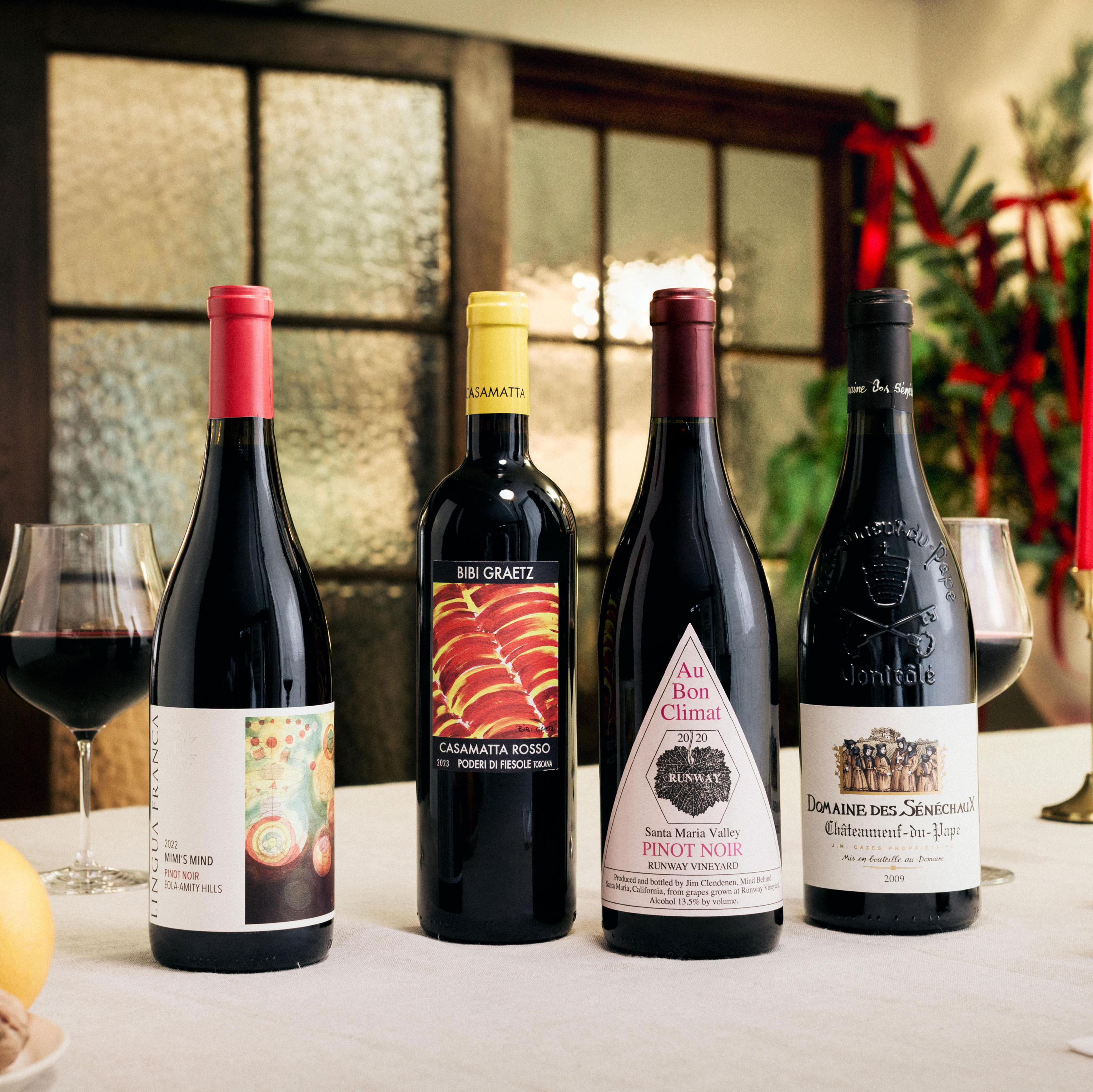 Four bottles of red wine are displayed on a festive dining table, accompanied by two glasses of red wine. The bottles feature labels with vibrant designs and colours, including red, yellow, and white. In the background, frosted glass doors, greenery with red ribbons, and tall red candles create a warm, elegant Christmas setting.