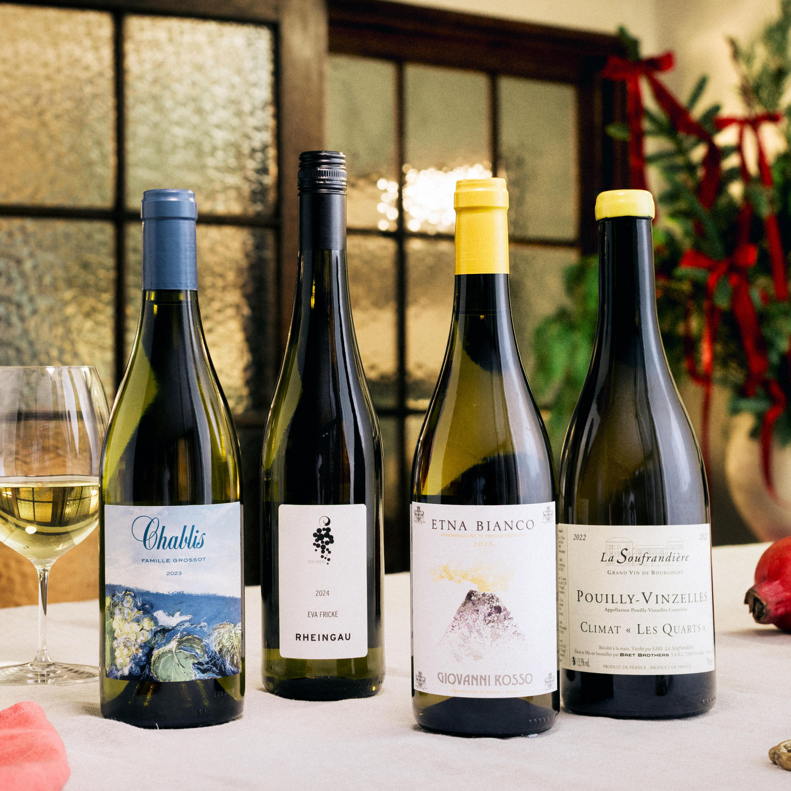 Four bottles of white wine are displayed on a festive dining table, accompanied by a glass of white wine. The bottles include Chablis, Rheingau, Etna Bianco, and Pouilly-Vinzelles, each with distinctive labels featuring blue, black, and yellow accents. The background features frosted glass doors, lush greenery with red ribbons, and soft, warm lighting, creating an elegant Christmas atmosphere.