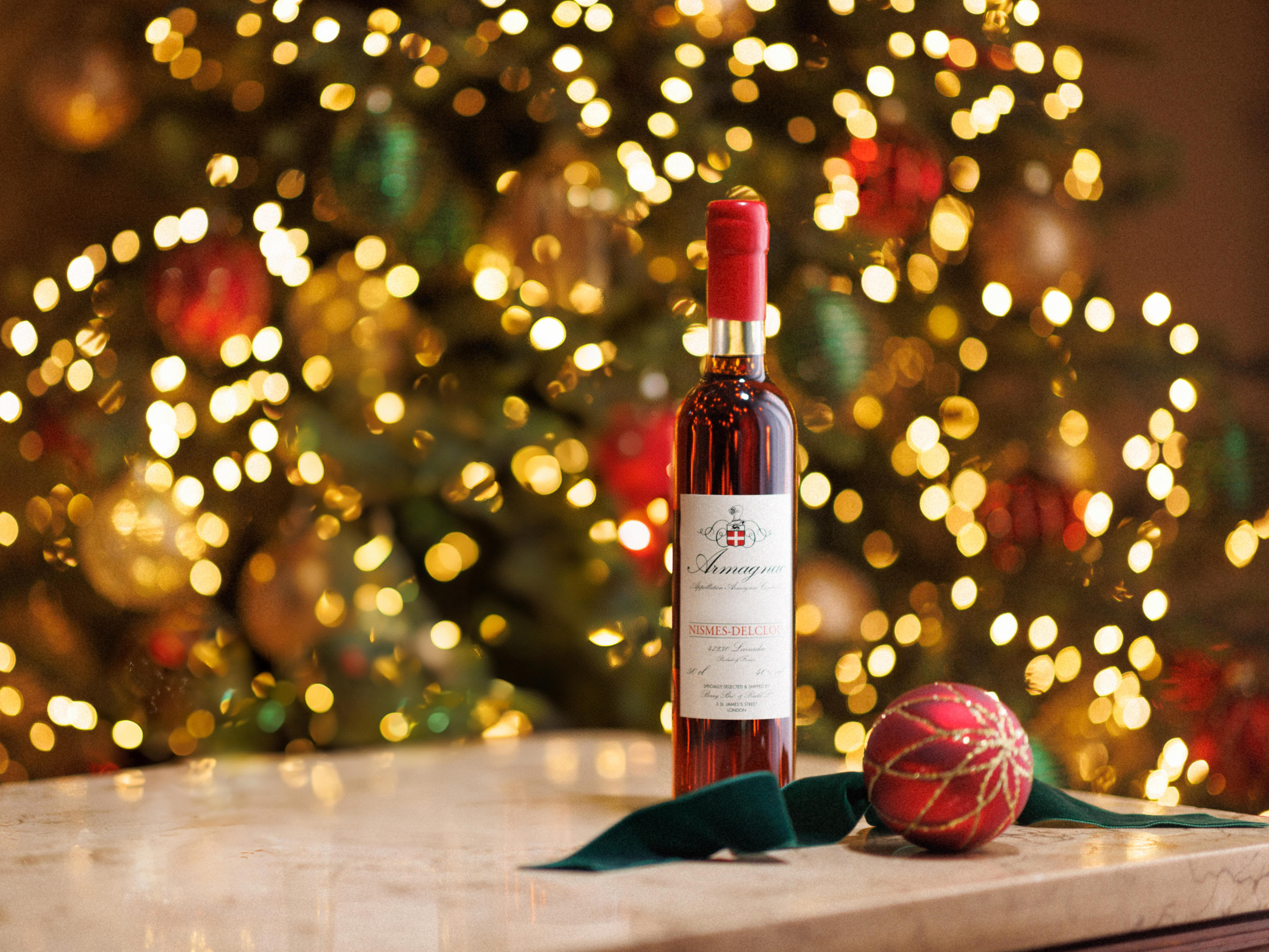A bottle of Armagnac spirit displayed on a festive surface with a red Christmas ornament and green cloth in the foreground. The background features a warmly lit Christmas tree with golden bokeh lights, creating a holiday atmosphere.