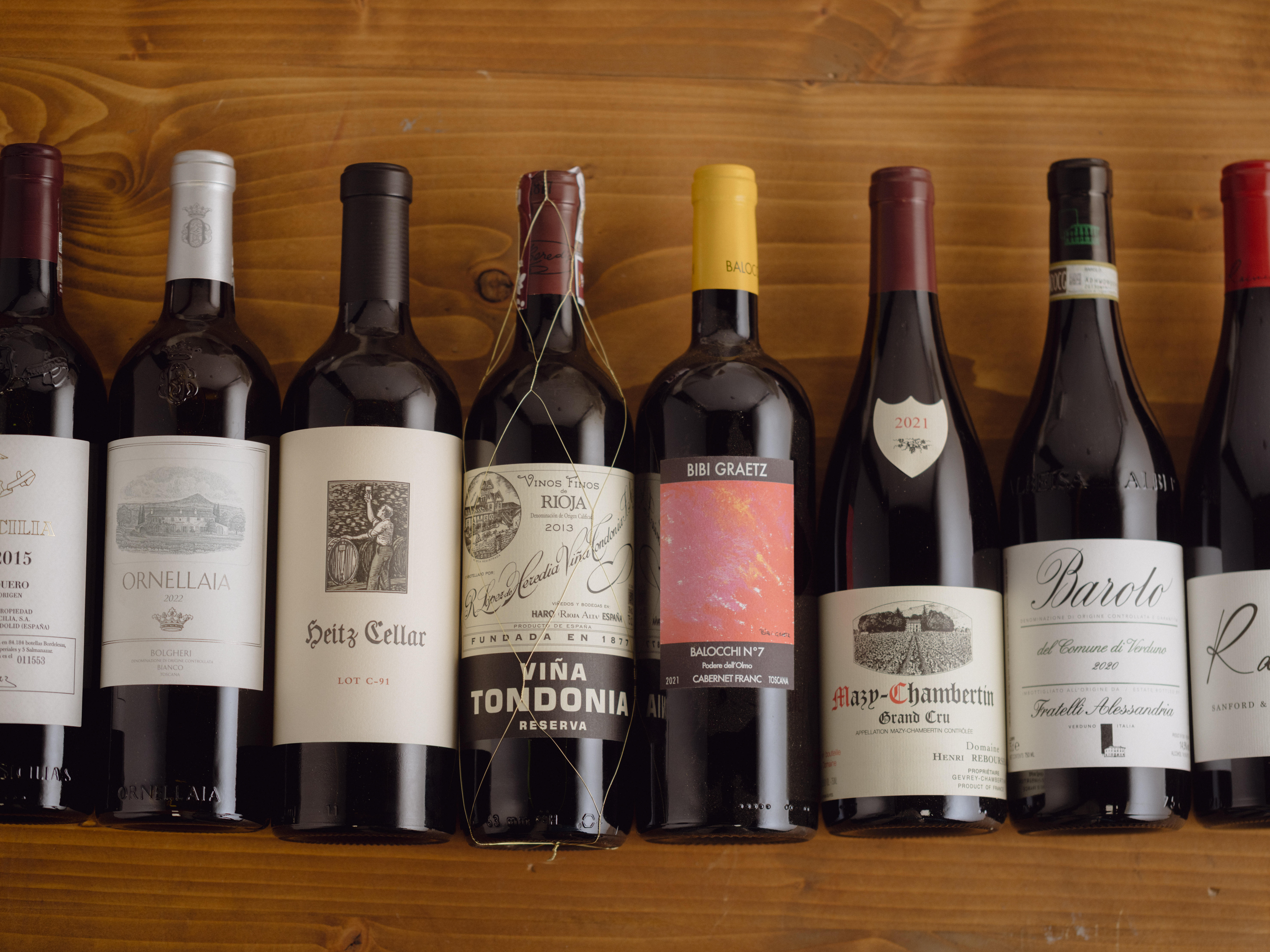 Eight assorted fine wine bottles on a wooden surface, including Ornellaia, Heitz Cellar, Viña Tondonia Reserva, Bibi Graetz Testamatta, Mazis-Chambertin Grand Cru, Barolo, and Ramey.