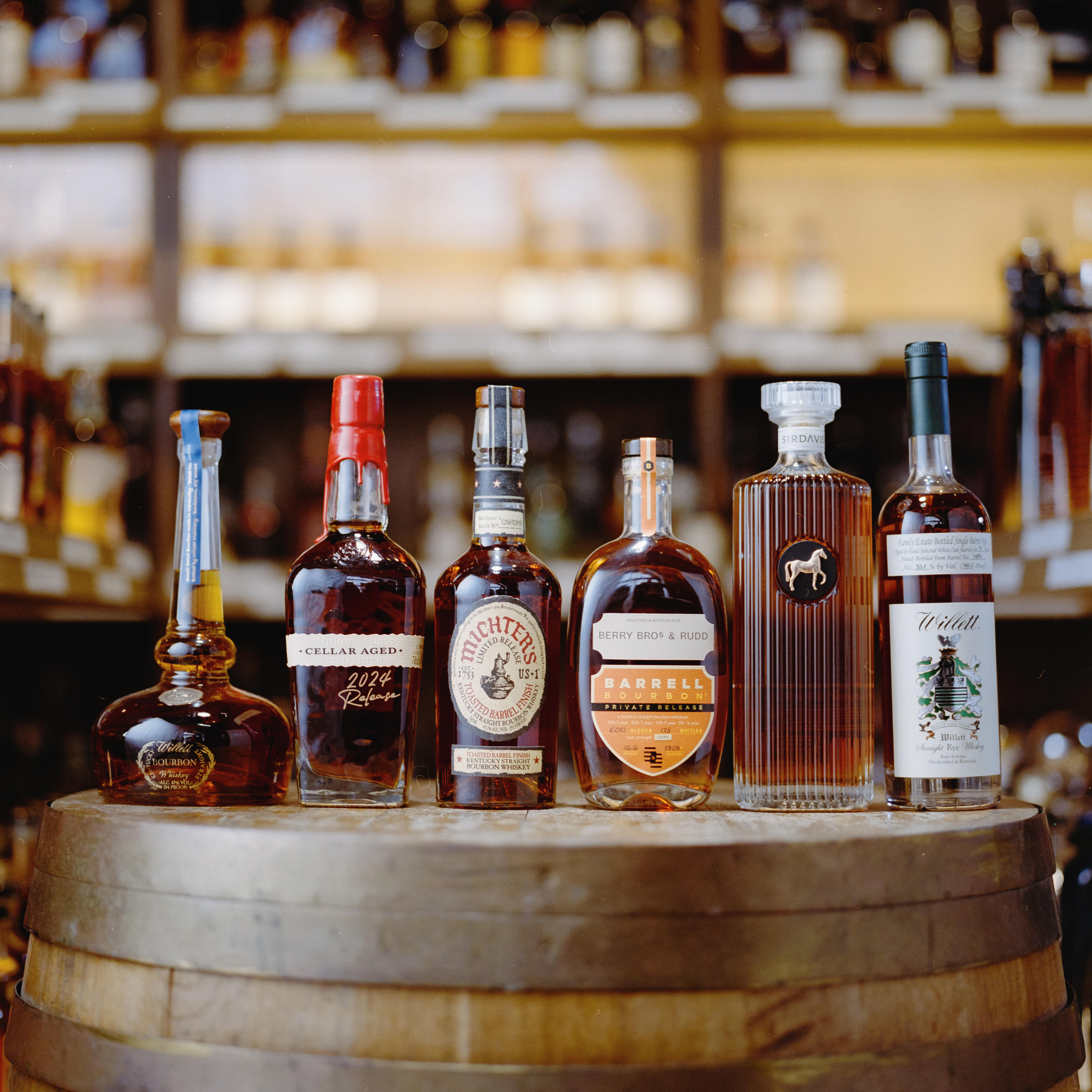 Film‑style close‑up (Portra 400 look) of Berry Bros. & Rudd’s bourbon and American whiskies collection. Several bourbon & US whisky bottles rest on a warm-toned surface with rich, moody lighting and fine film grain textures, conveying a classic, cinematic and premium mood.