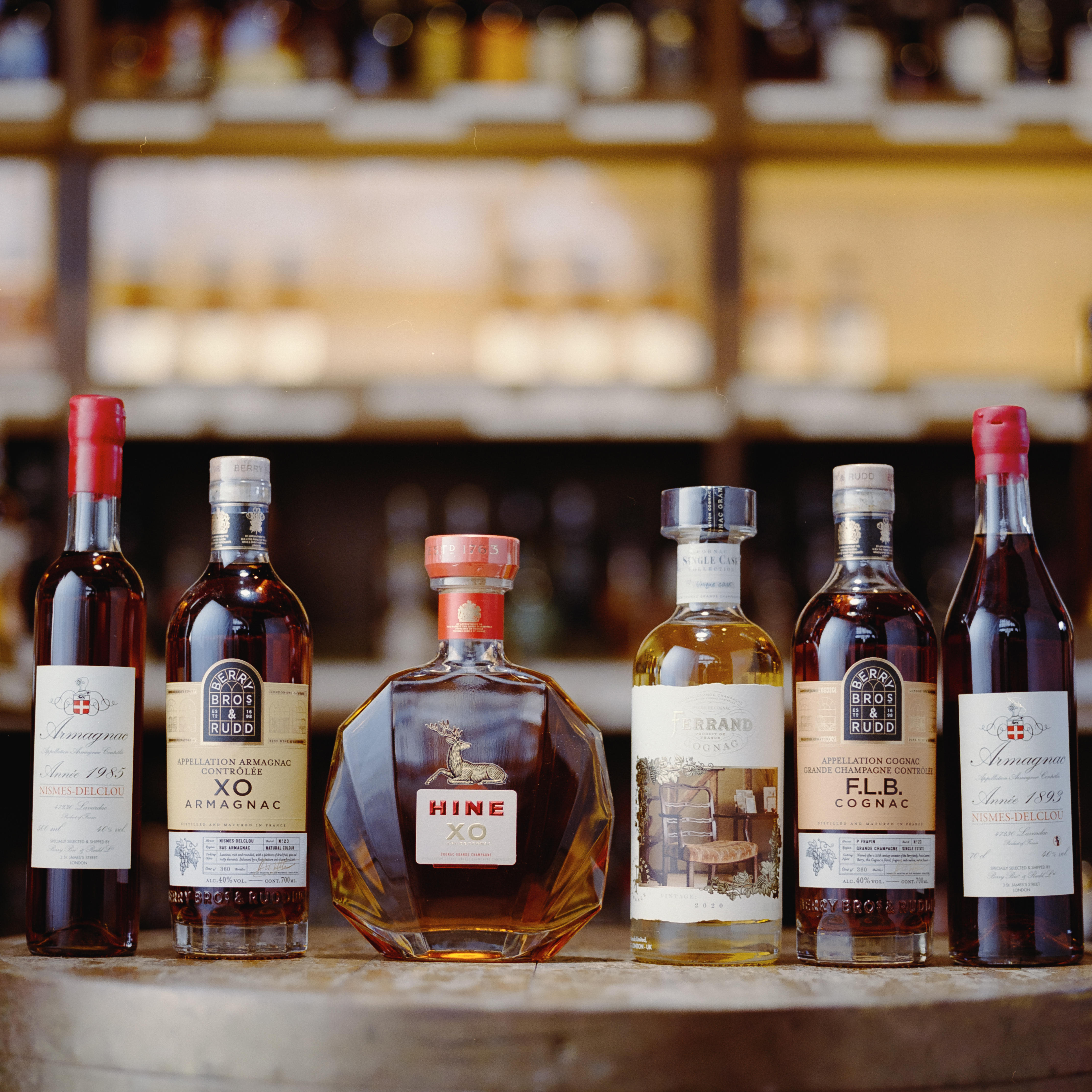 A selection of premium Cognac and Armagnac bottles, including Hine XO, Ferrand, and Berry Bros. & Rudd own-label, displayed on a wooden cask with spirits shelves softly blurred behind.