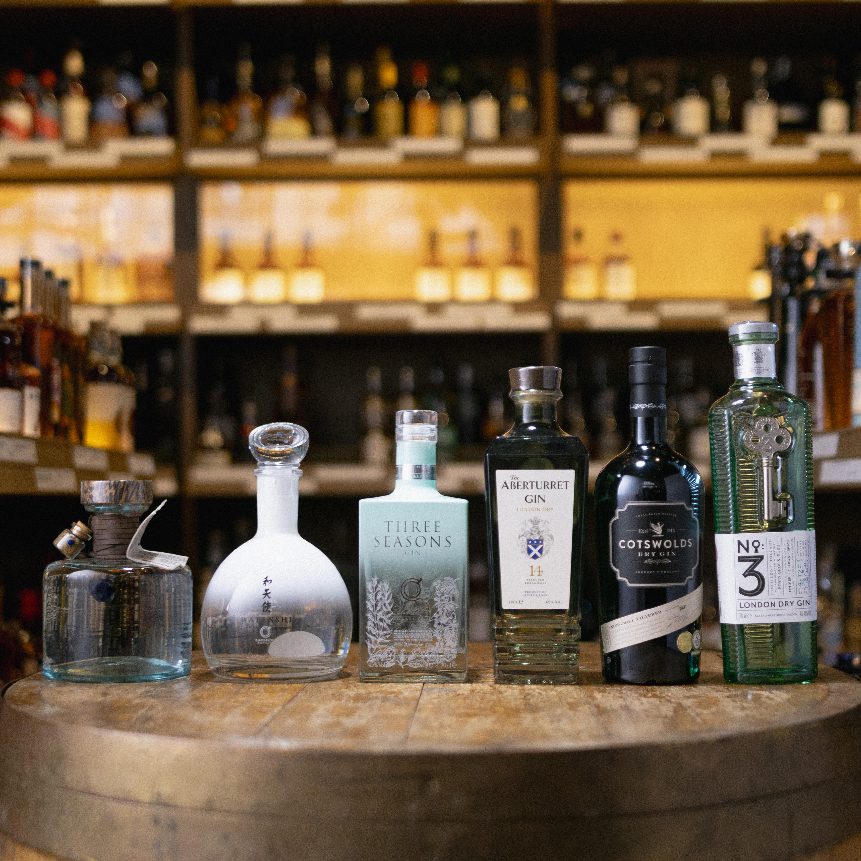 A selection of premium gins, including No.3, Cotswolds, Aberturret and Three Seasons, displayed on a wooden cask with spirits shelves blurred in the background.