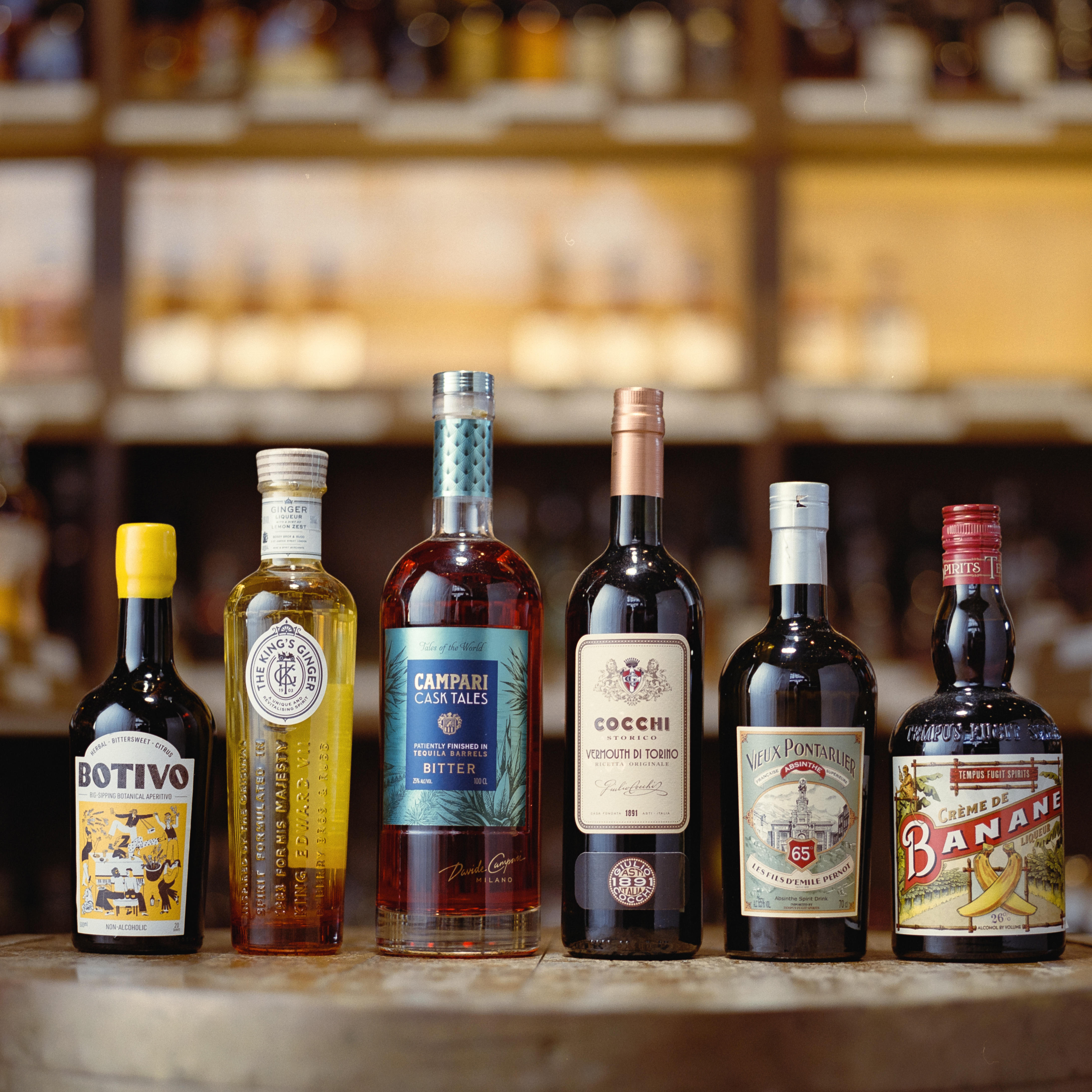 A selection of aperitifs and cocktail ingredients is displayed on a wooden cask, featuring brands like Campari, Cocchi, Vieux Pontarlier, Crème de Banane, and Botivo. Softly blurred spirits shelves are in the background.