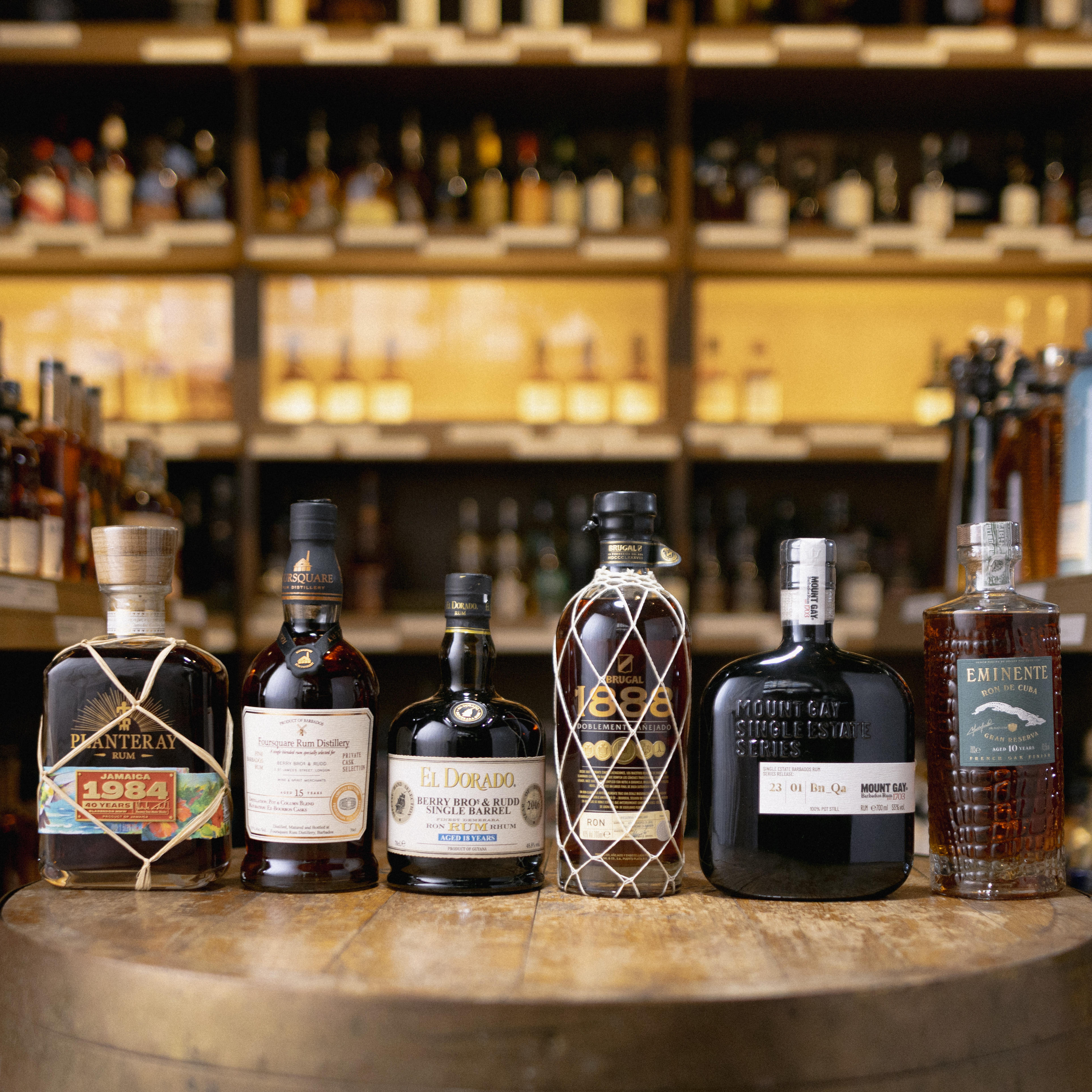 A selection of premium rums on a wooden cask features brands like Plantation, El Dorado, Brugal, Mount Gay, and Eminente, with blurred spirits shelves in the background.