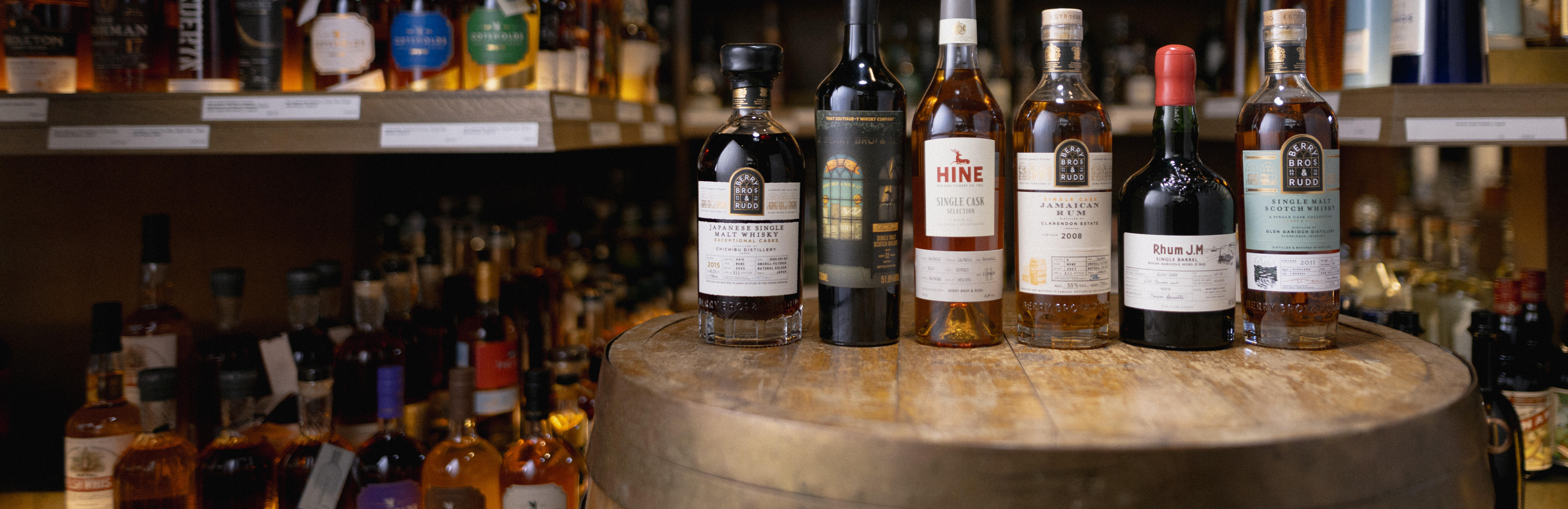 A selection of Berry Bros. & Rudd's own-label spirits displayed on a wooden cask: Japanese whisky, Scotch, Jamaican rum, Cognac, and other limited-edition bottlings, with shelves of premium spirits blurred in the background.