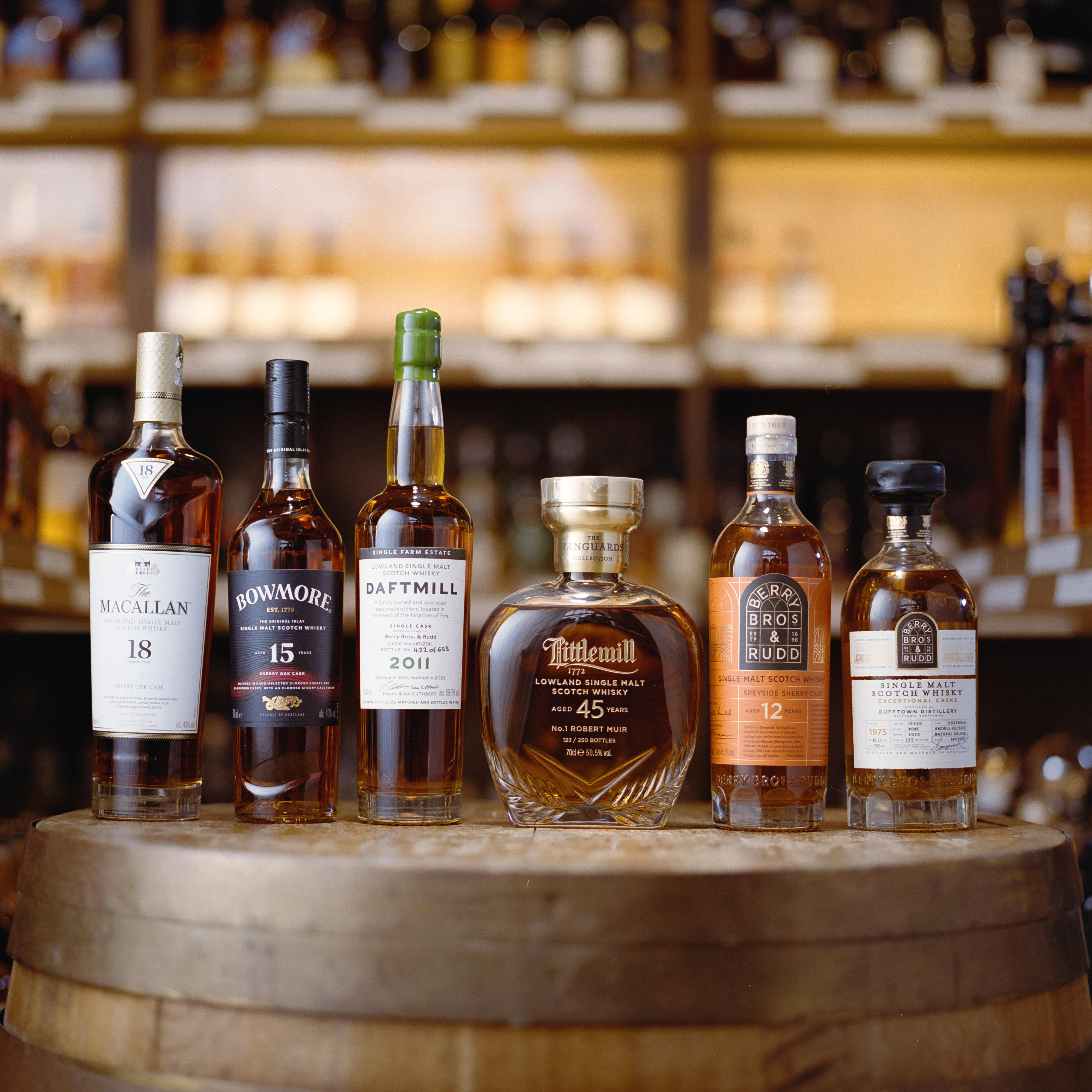 Close-up film-style shot (Portra 400 aesthetic) showcasing a range of Berry Bros. & Rudd scotch whiskies. Several whisky bottles are positioned on a warm-toned surface with rich, moody lighting and subtle film grain. The photo evokes a classic, cinematic mood focused on premium spirits.