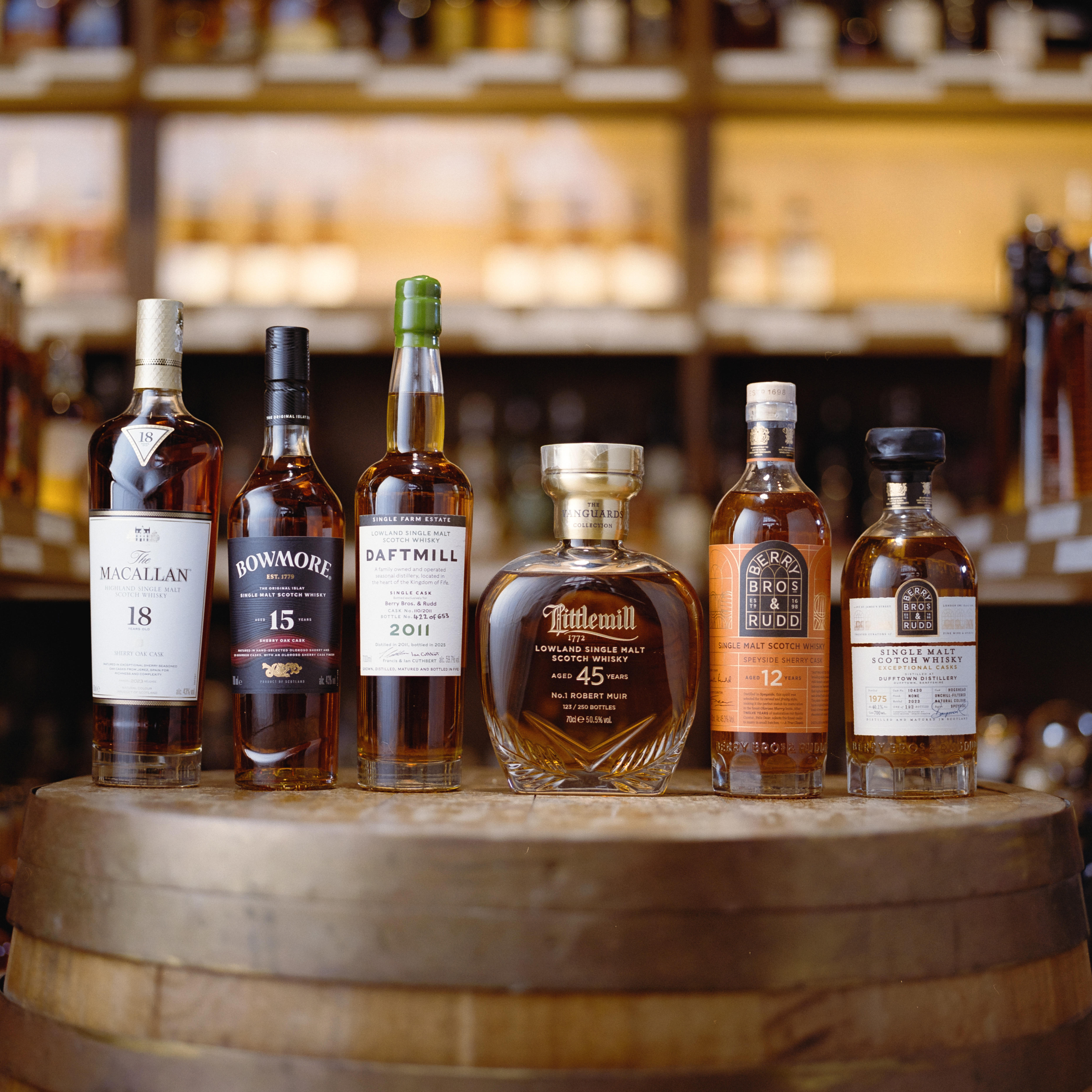 Six premium Scotch whiskies on a wooden barrel at Berry Bros & Rudd, including The Macallan 18, Bowmore 15, Daftmill 2011, Tittlewell 45, and Edradour 12, with shelves of fine spirits in the background.