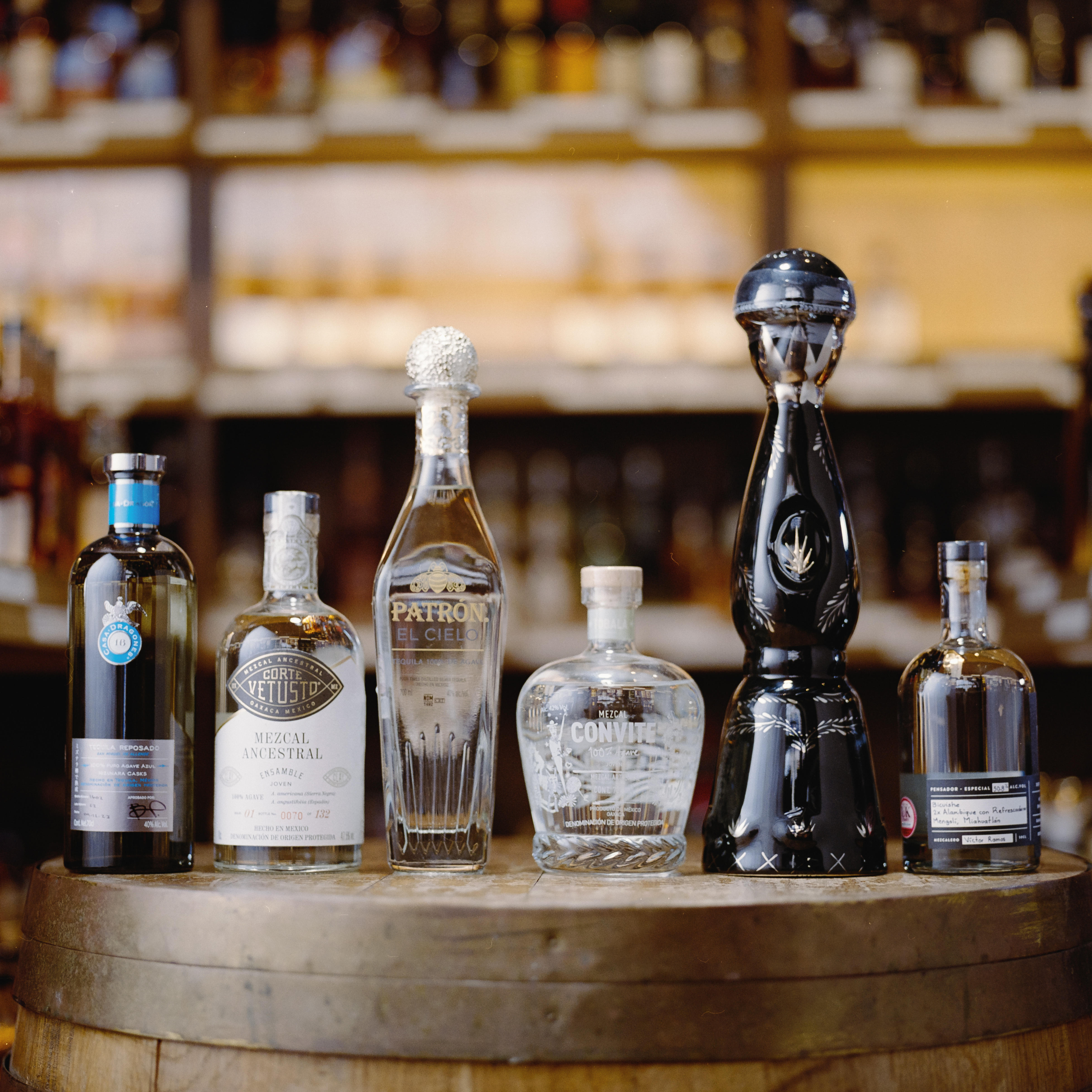 Selection of premium Tequilas and Mezcals on a wooden cask, including Patrón El Cielo, Clase Azul, Convite and Corte Vetusto, with spirits shelves softly blurred in the background.