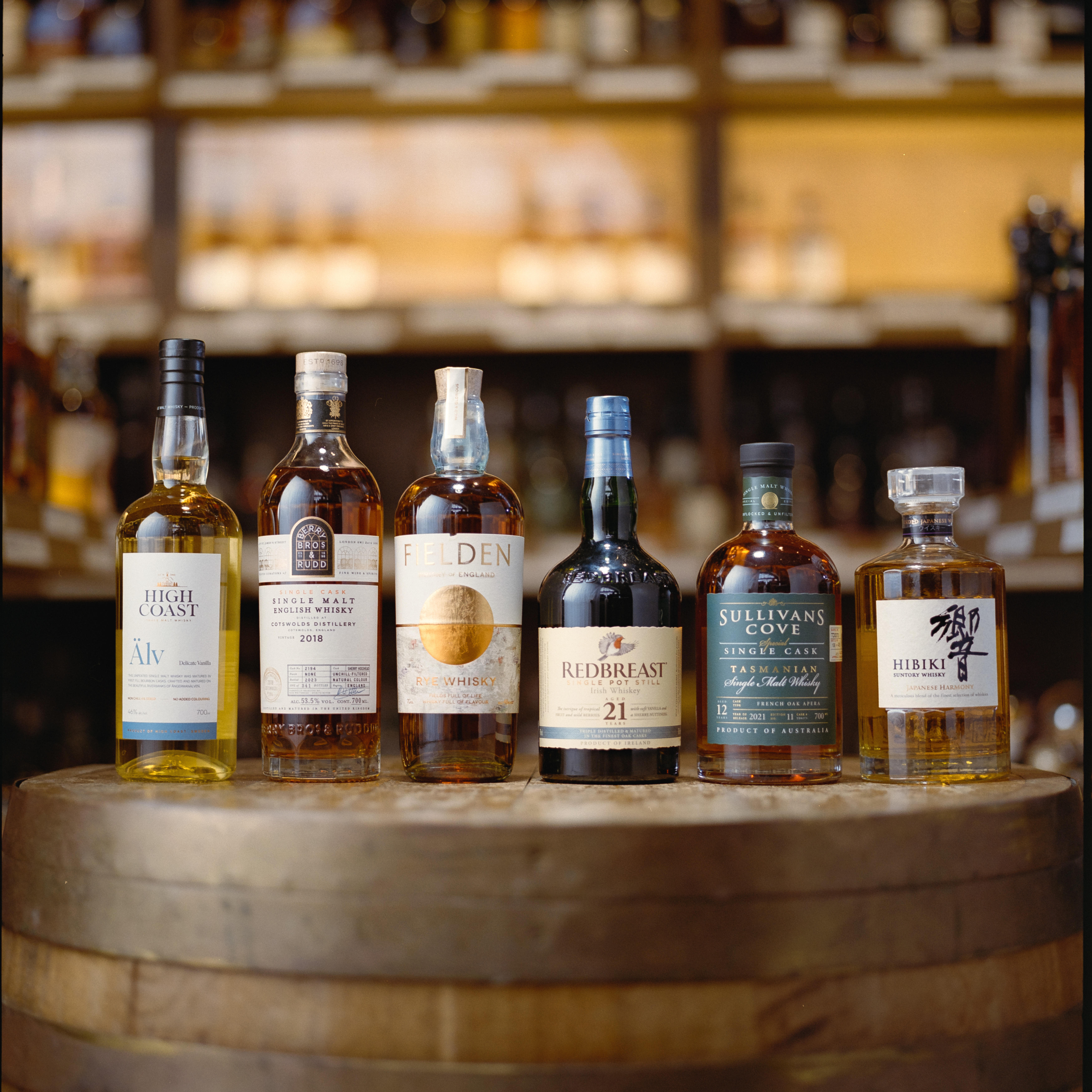 Film‑style close‑up (Portra 400 look) capturing Berry Bros. & Rudd’s “Whiskies of the World” selection: an assortment of international whisky bottles (Scotch, American, Japanese, Irish) arranged on a warm-toned surface under moody, cinematic lighting with subtle film grain for texture.