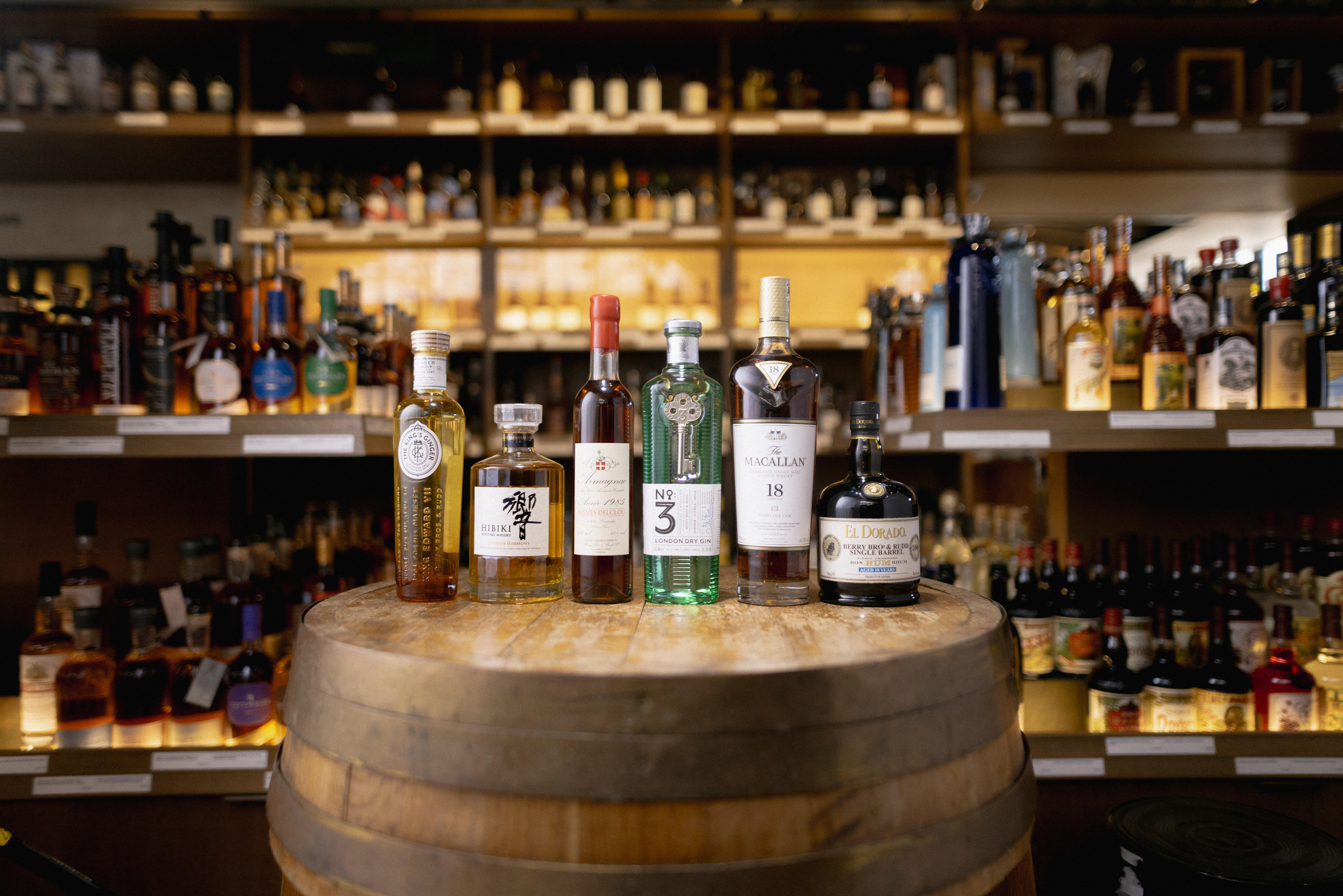 A behind‑the‑scenes group shot of Berry Bros. & Rudd’s spirits line‑up: a variety of premium whisky, rum and gin bottles artfully arranged on a table with soft studio lighting